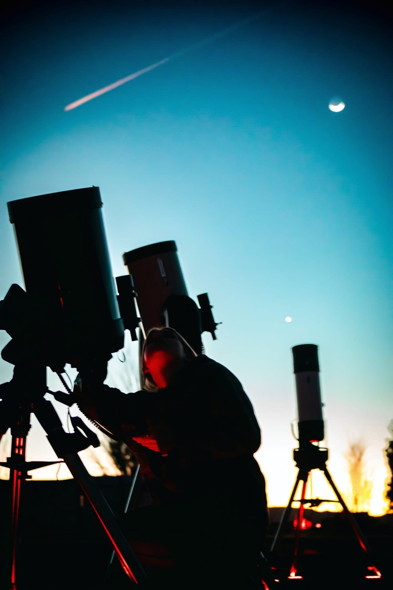 A guide adjusting a telescope at dusk, with a crescent moon and a bright planet overhead