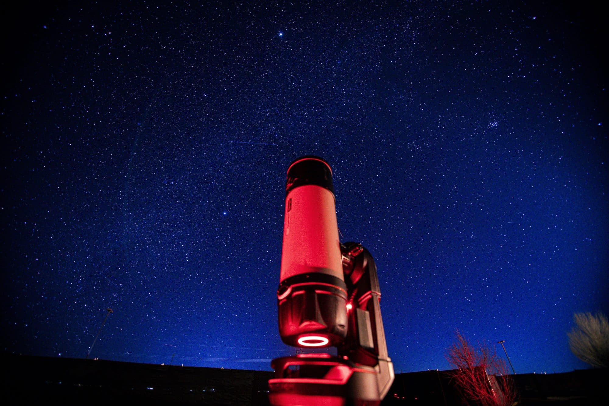 A single telescope in red light under a deep blue sky thick with stars