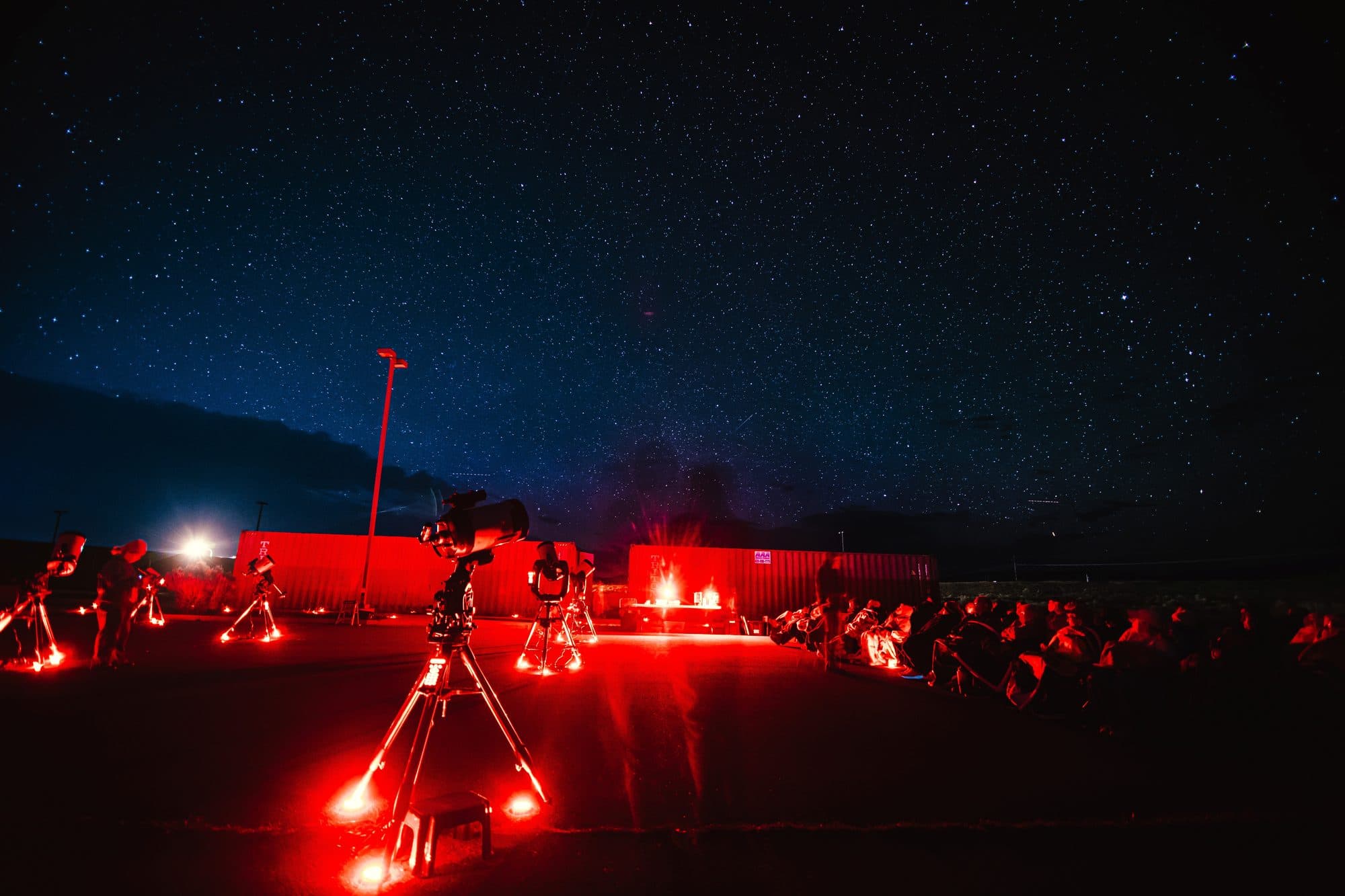 A row of telescopes set up facing a seated group under the night sky