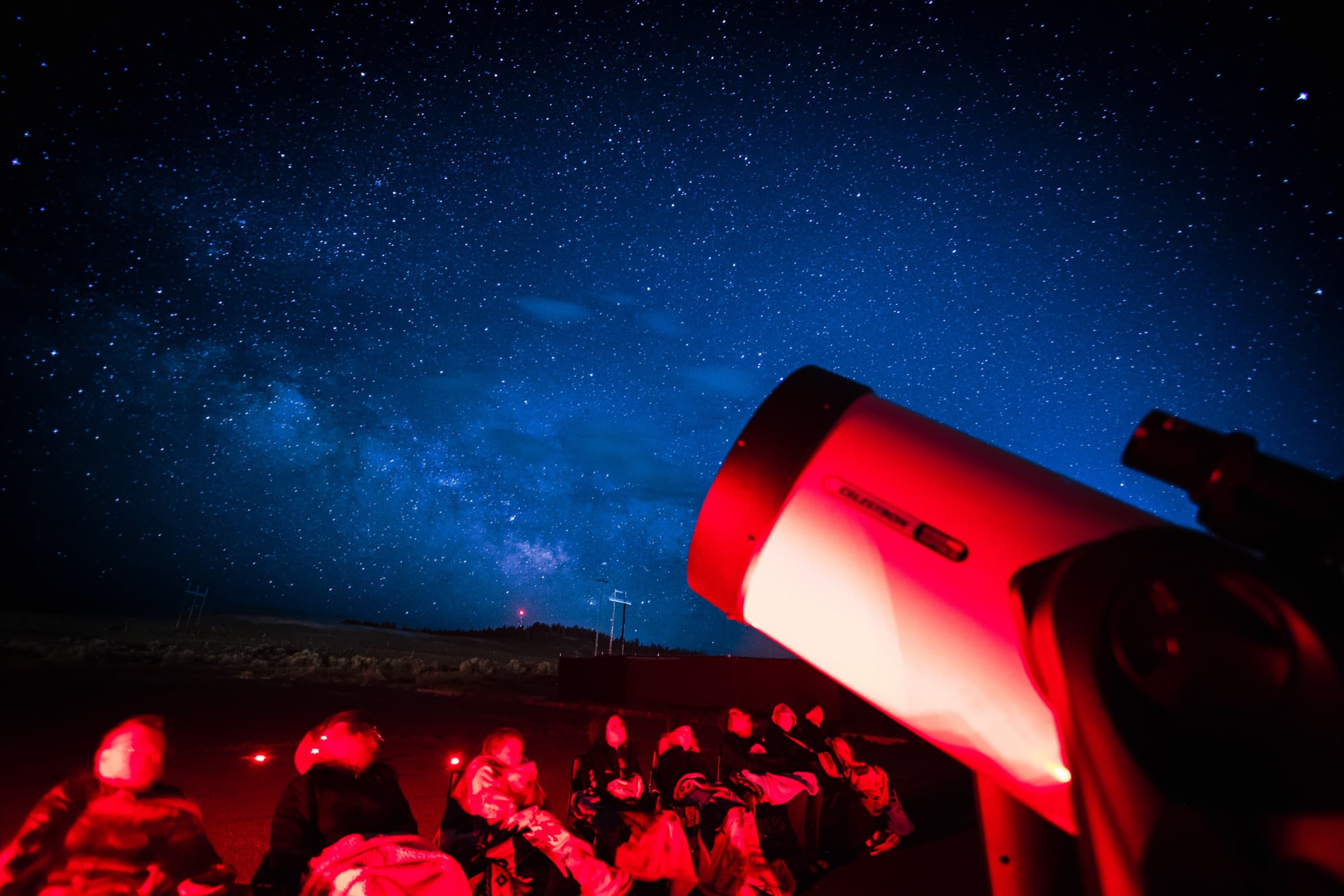 Guests wrapped in blankets watching from their chairs as a large telescope points into the Milky Way