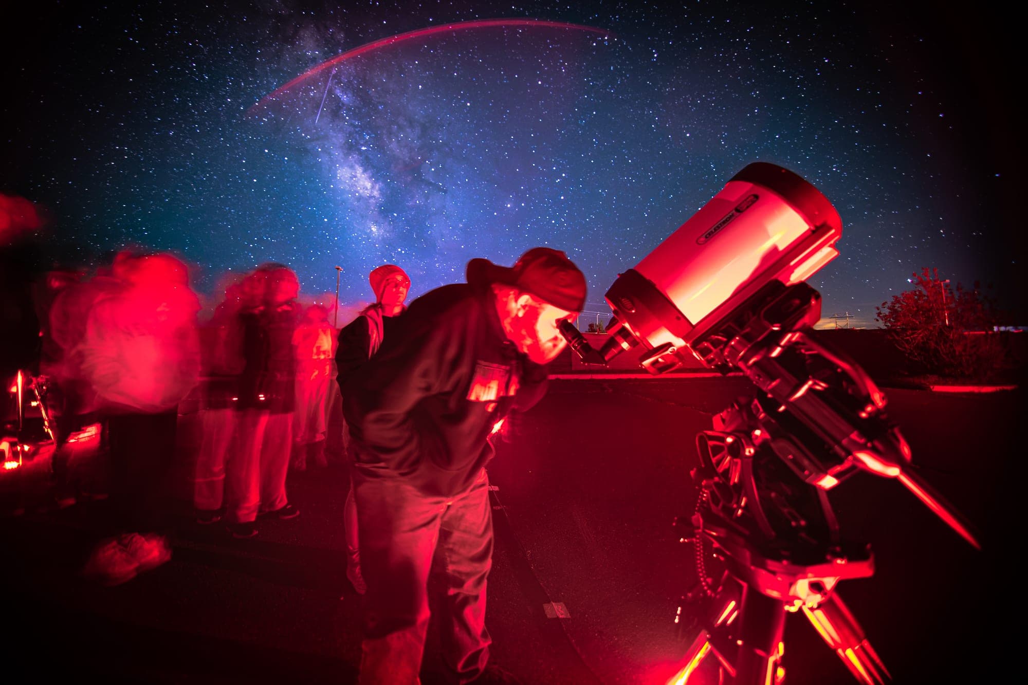 A guide leaning into the eyepiece in red light, the Milky Way arching behind him