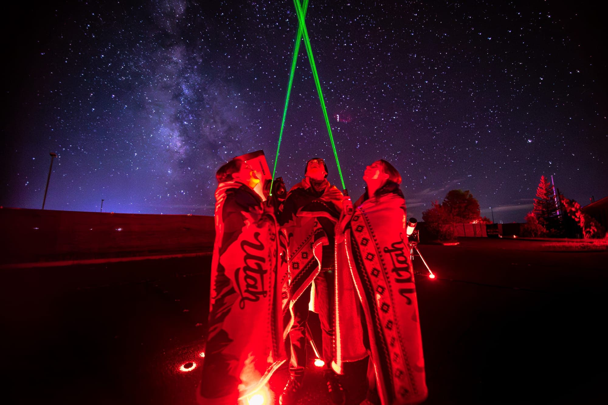 Three guests wrapped in Utah blankets as two green lasers cross the Milky Way overhead