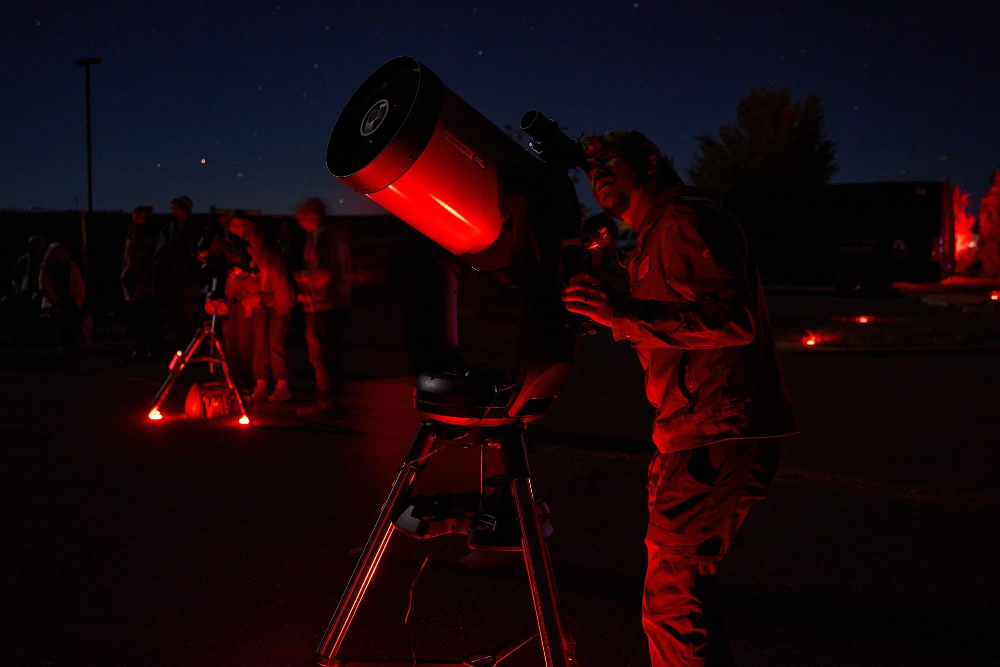 A guide at the eyepiece of a large telescope in full red light, guests waiting behind